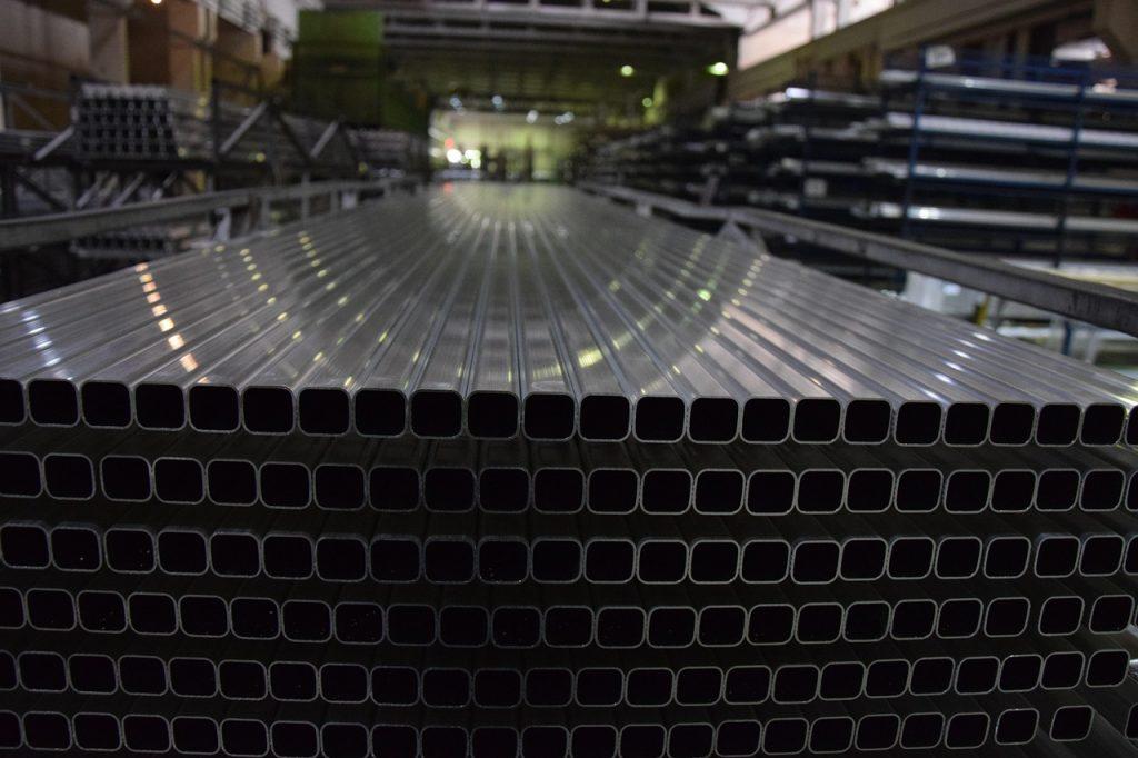 Perfiles cuadrados de aluminio recién extruidos apilados ordenadamente en el interior de una gran nave industrial.