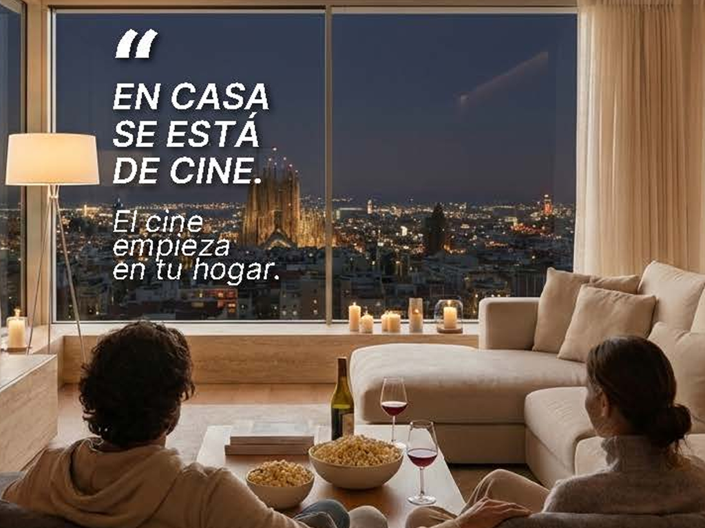 Pareja en un salón moderno con grandes ventanales de aluminio viendo la ciudad de Barcelona de noche, con el texto "En casa se está de cine".