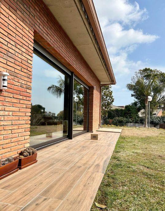 Perspectiva del porche de una casa con fachada de ladrillo y amplios ventanales correderos que reflejan el entorno natural.