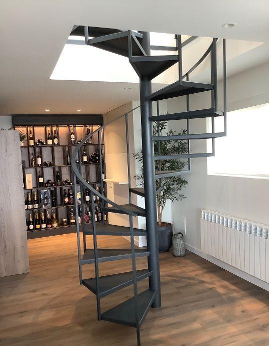 Escalera de caracol metálica oscura en un salón moderno con suelo de madera, un gran mueble botellero al fondo y una ventana de aluminio a la derecha.