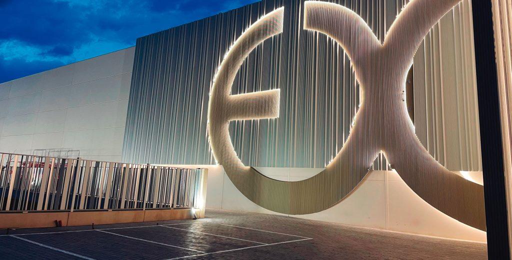 Fachada industrial nocturna con un gran logotipo "EX" iluminado y revestimiento de lamas de aluminio.