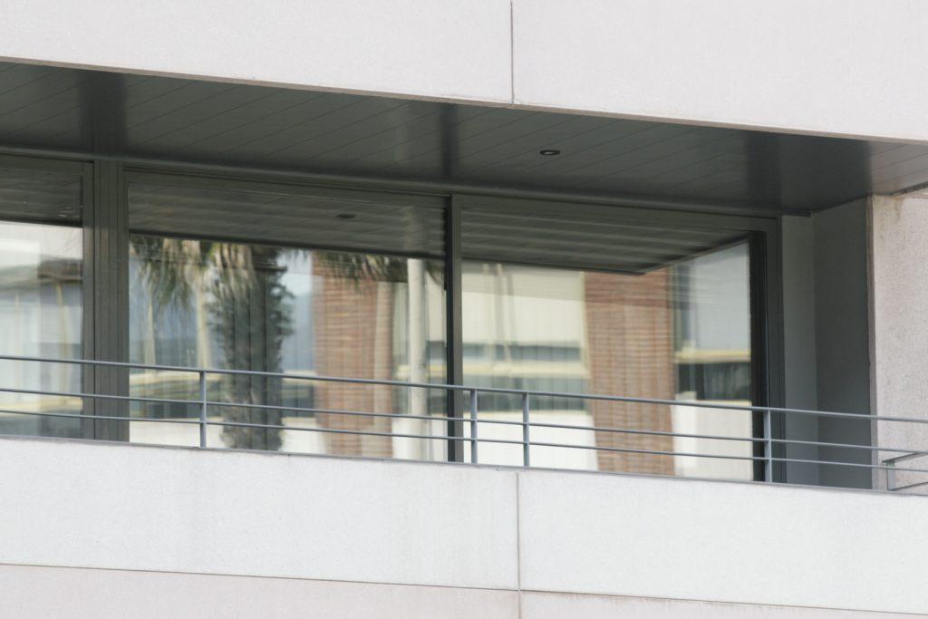 Detalle exterior de una ventana y puerta corredera de aluminio bajo un falso techo de lamas de aluminio oscuro Apotec.
