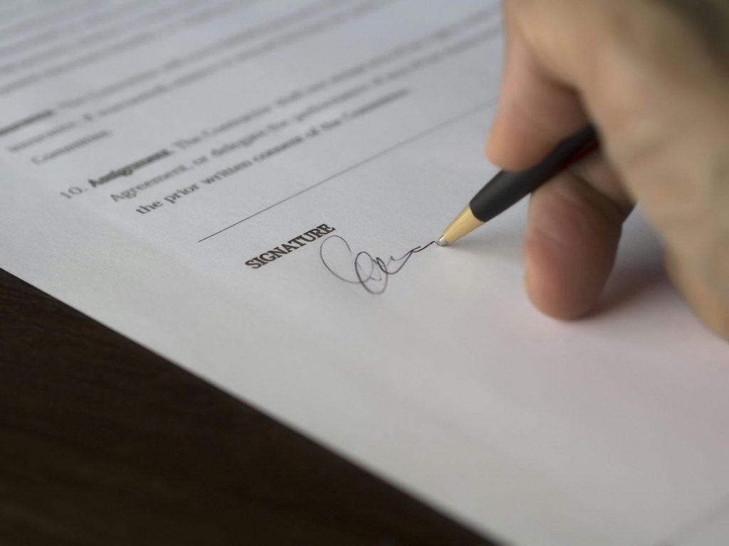 Mano sosteniendo un bolígrafo firmando un documento o contrato sobre la línea de "Signature".