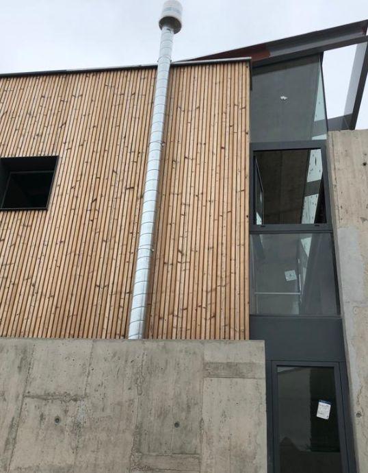 Vista exterior de una vivienda con muro de hormigón, fachada de madera, un tubo metálico y ventanales estrechos de aluminio.