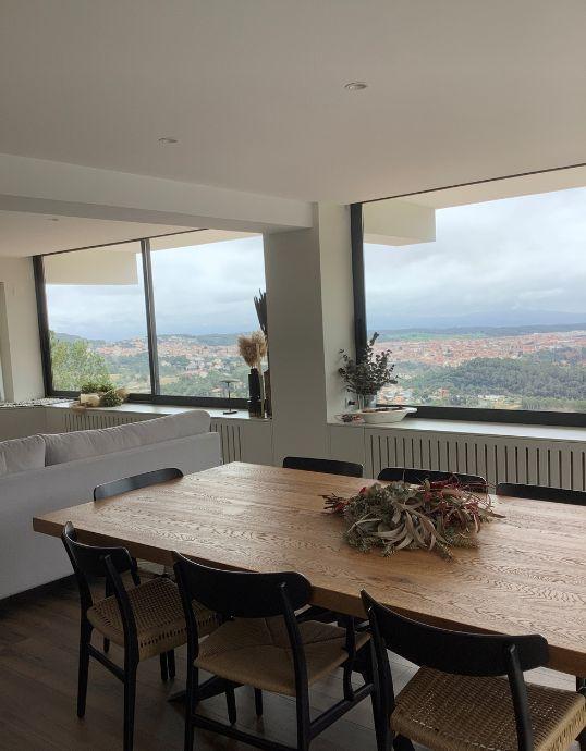 Perspectiva del salón de la Casa Metaldos mostrando la mesa del comedor y grandes ventanales de aluminio instalados sobre muebles blancos cubrerradiadores con vistas al pueblo y la montaña.
