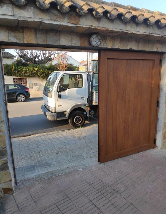 Puerta de garaje de aluminio efecto madera parcialmente abierta mostrando la calle y un camión en Casa en Vilassar de Dalt.