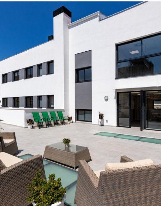 Amplia terraza amueblada frente a la fachada blanca de un edificio con ventanas rectangulares y puertas de acceso de aluminio oscuro en Badalona.
