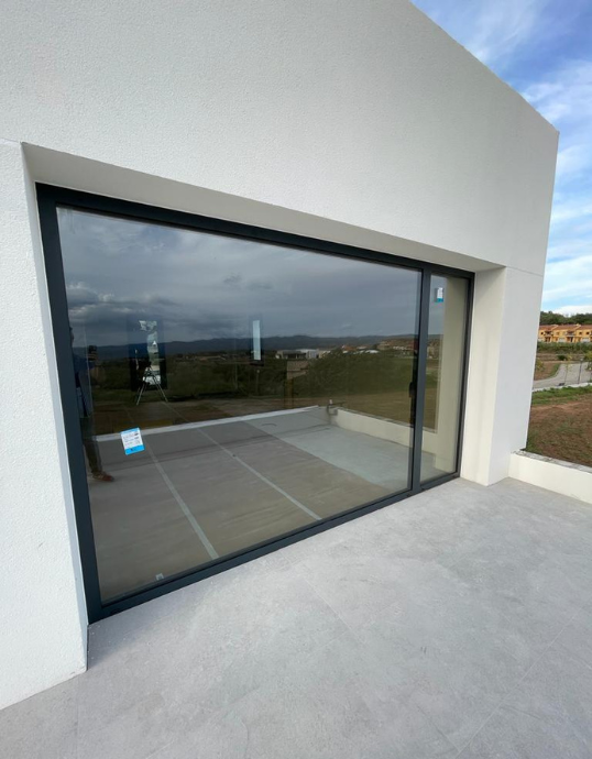 Gran ventanal de cristal con perfilería de aluminio oscuro instalado en una fachada blanca de diseño moderno en la Casa en Sant Feliu Saserra.