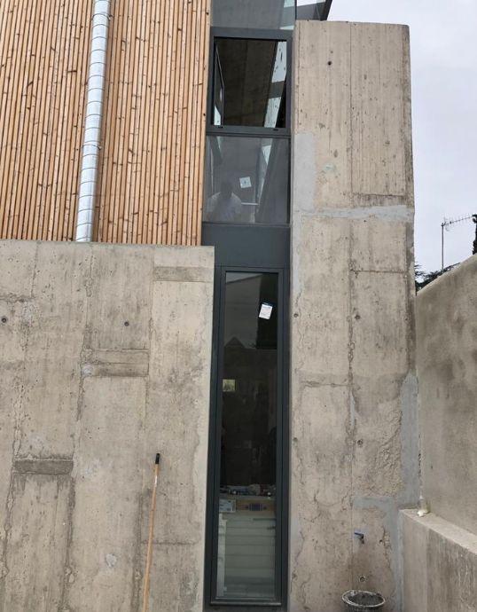 Ventanal estrecho y vertical de aluminio oscuro instalado entre un muro de hormigón y una fachada de madera.