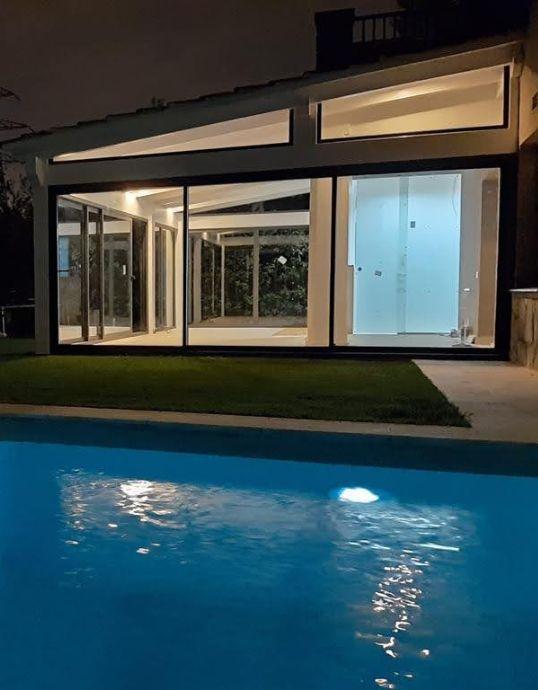 Vista nocturna de una casa moderna con piscina iluminada y grandes ventanales correderos de aluminio oscuro en la fachada.