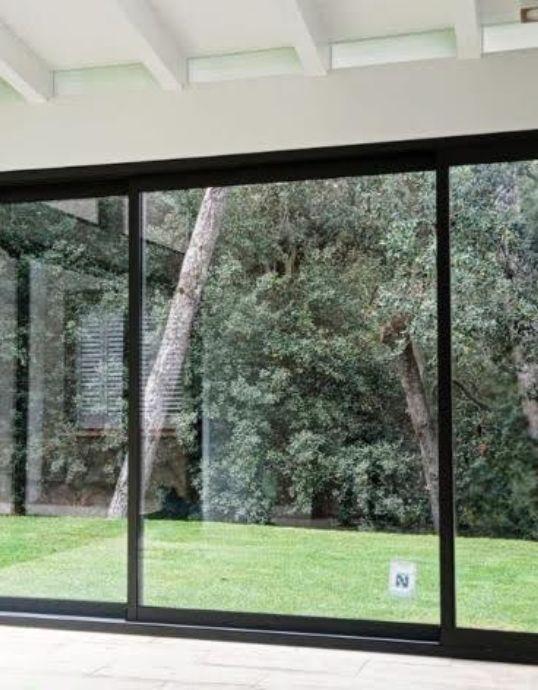 Grandes ventanales correderos de aluminio negro que enmarcan un frondoso jardín con árboles altos desde el interior de una casa con techo de vigas blancas.